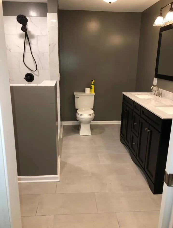 Full bathroom remodel with a walk-in tile shower with two shower heads, a dark shaker double vanity with marble-look tops, painted gray-green walls and large-format floor tile