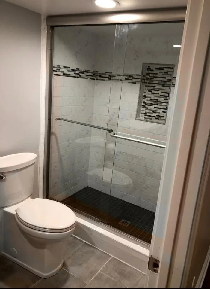 Remodeled bathroom with a sliding glass shower door, marble-look wall tile, a dark glass mosaic accent band and matching recessed niche, a dark tile shower floor and a new toilet