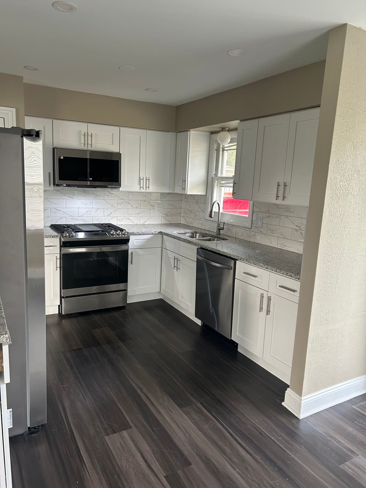 Remodeled kitchen with white shaker cabinets, speckled gray granite countertops, a marble-look plank tile backsplash, stainless steel range, microwave, dishwasher and refrigerator, and dark wood-look plank flooring