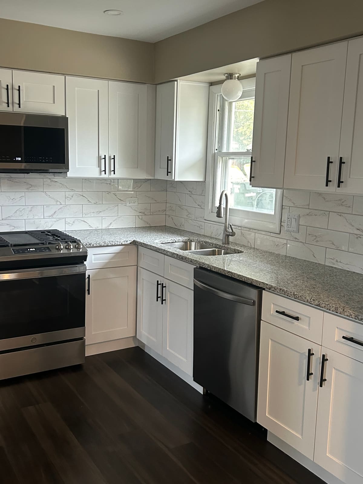 Corner of a remodeled kitchen with white shaker cabinets and black bar pulls, speckled granite countertops, a marble-look tile backsplash, an undermount double sink with a black gooseneck faucet under the window, and a stainless steel range and dishwasher