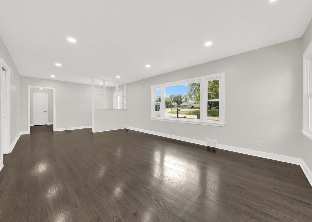 Empty living room finished with dark stained hardwood flooring, light gray walls, white baseboards and window casing, recessed ceiling lights, a wide white-trimmed picture window and a white built-in shelving partition