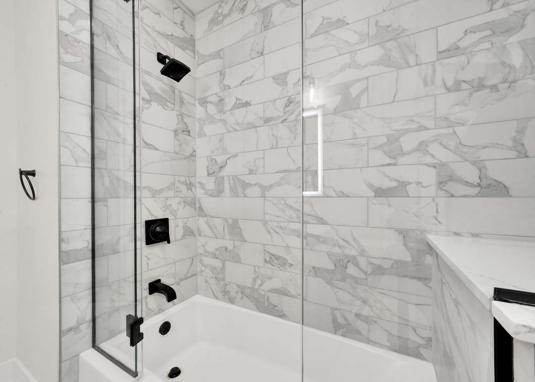 Tub and shower surround tiled floor to ceiling in large-format marble-look porcelain, with a matte black shower head, valve trim and tub spout, a black-framed glass shower screen and a white tub