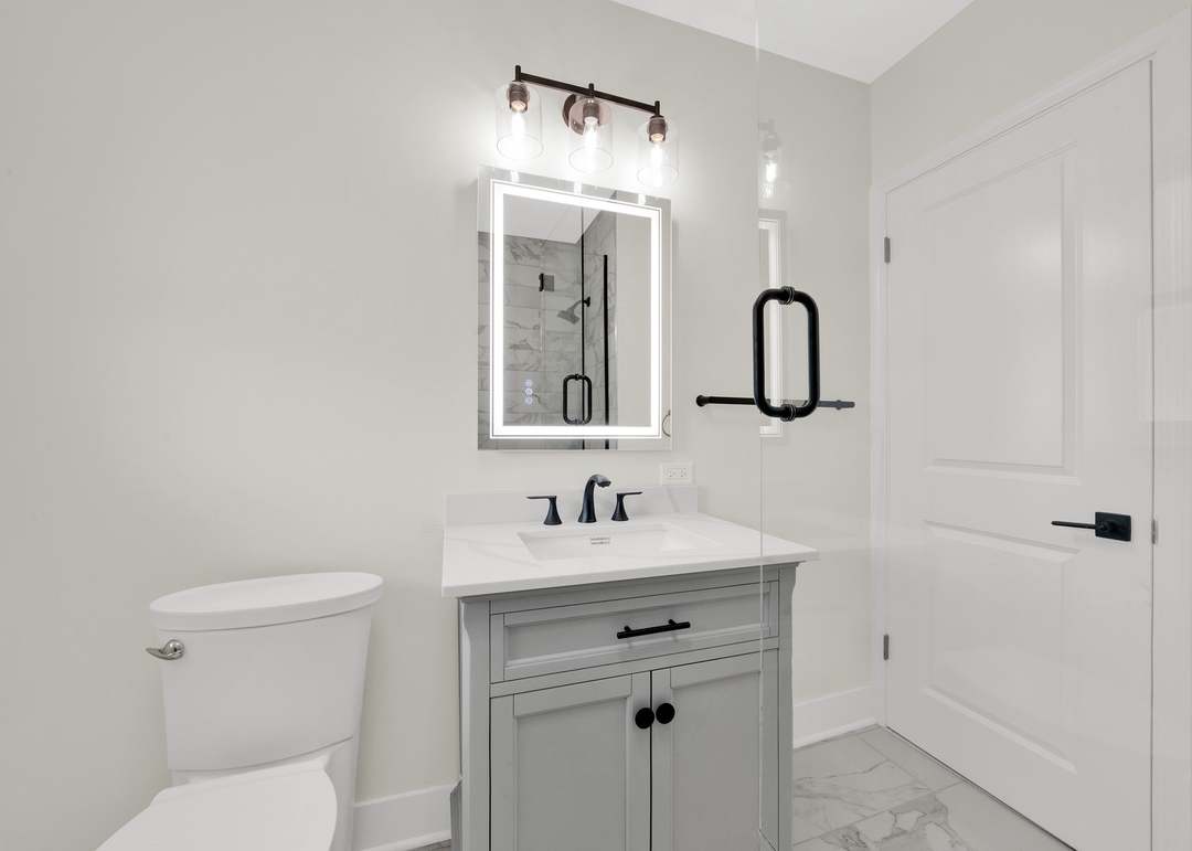 Remodeled bathroom with a gray shaker vanity and white stone-look top, a matte black widespread faucet, a backlit LED mirror, a three-light vanity fixture, a black towel bar and marble-look floor tile