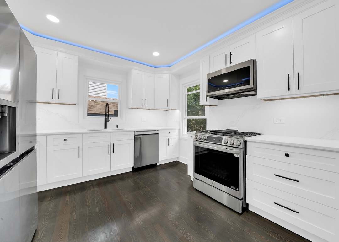 Remodeled kitchen with white shaker cabinets and black bar pulls, white quartz countertops and a full-height stone-look backsplash, stainless steel range, microwave, dishwasher and refrigerator, blue LED cove lighting in the ceiling and dark stained wood flooring