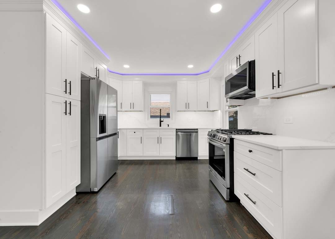 Galley kitchen with white shaker cabinets to the ceiling, black bar pulls, white quartz countertops and stone-look backsplash, stainless steel refrigerator, gas range, over-range microwave and dishwasher, purple LED cove lighting around the dropped ceiling and dark stained wood flooring