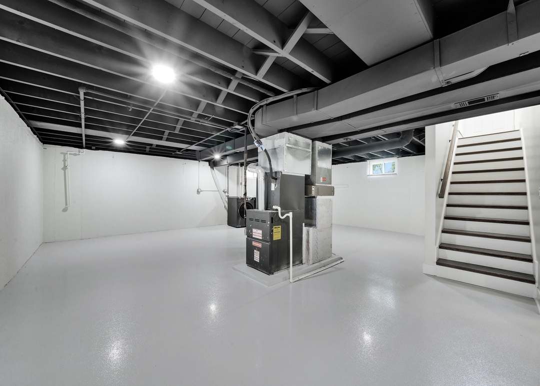 Cleaned-up basement with white painted walls, a light gray coated concrete floor, black painted exposed joists and ductwork, a furnace and a staircase with dark treads and white risers
