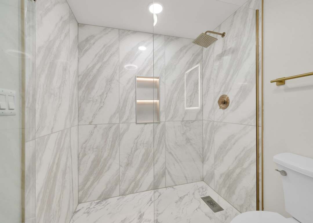 Walk-in shower tiled floor to ceiling in large-format marble-look porcelain, with a brushed gold rain shower head and valve trim, a lit two-shelf recessed niche, a gold-framed glass shower screen and a marble-look shower floor with a square drain