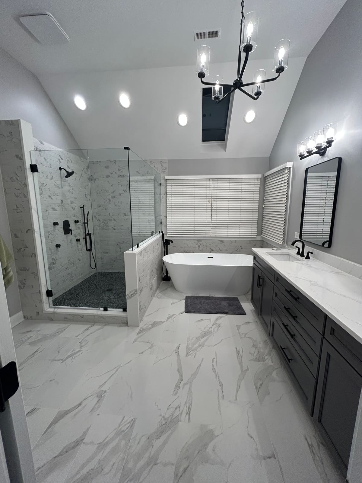 Vaulted bathroom with a freestanding white soaking tub and matte black floor-mounted tub filler, a glass corner shower tiled in marble-look porcelain over a dark pebble shower floor, a dark gray vanity with a marble-look quartz top, a black-framed mirror and black chandelier, over large-format marble-look floor tile