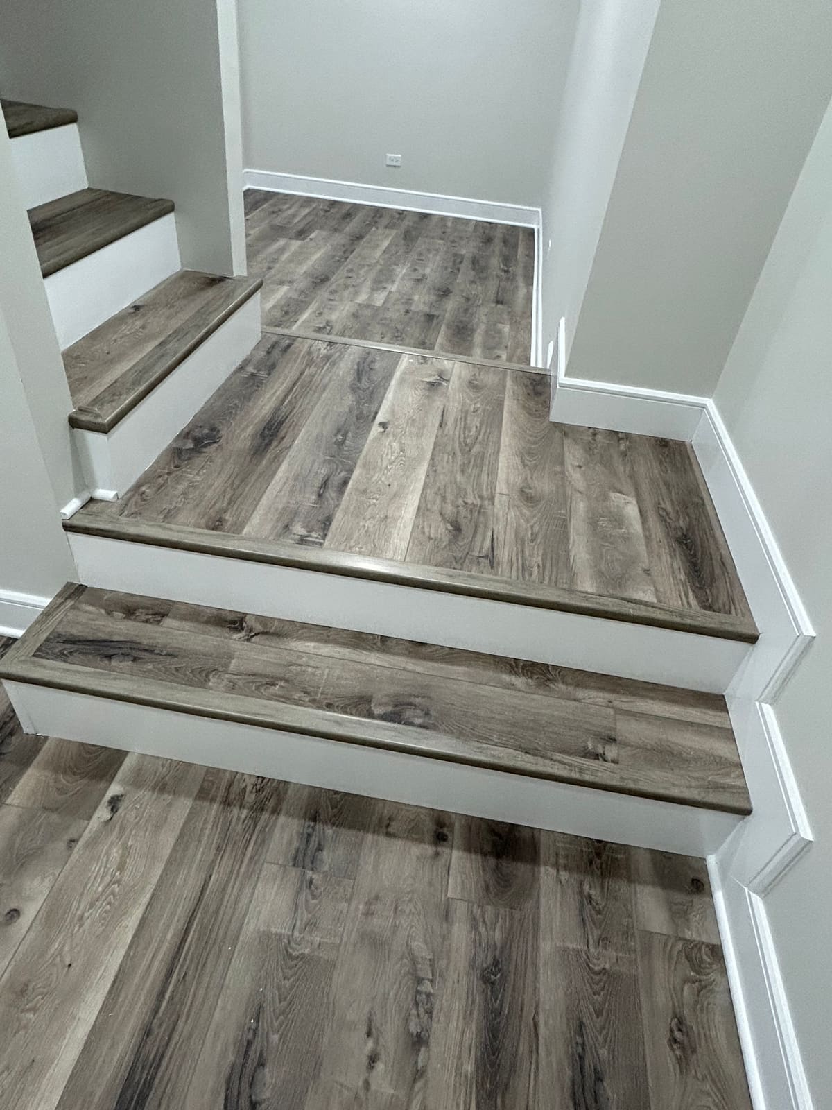 Staircase landing finished with gray-brown wood-look plank treads and freshly painted white risers, skirt board and baseboard, opening onto a hallway laid with matching plank flooring