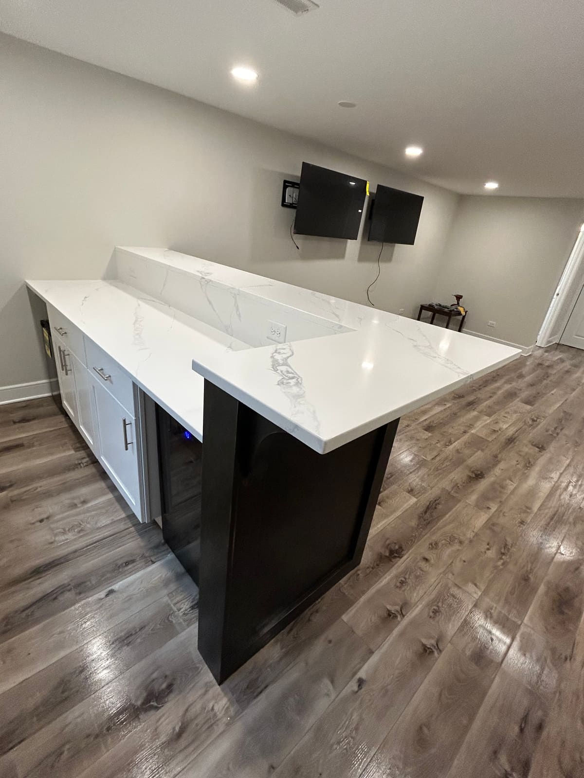 Finished basement bar with a white marble-look quartz countertop and raised bar top, white shaker base cabinets with brushed nickel bar pulls, a dark espresso bar front, an under-counter beverage refrigerator, recessed ceiling lights, two wall-mounted flat-screen televisions and gray-brown wood-look plank flooring