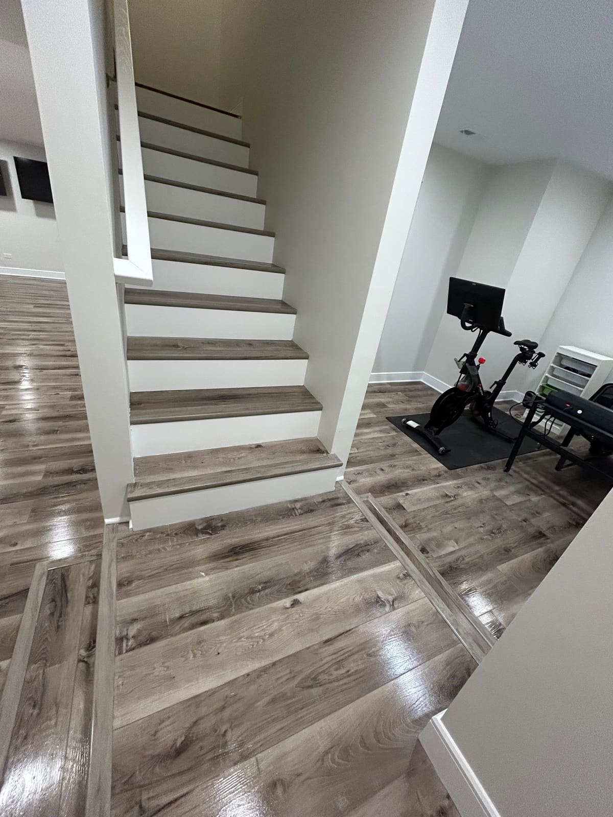 Finished basement staircase with gray-brown wood-look plank treads, freshly painted white risers, skirt board and wrapped support posts, a wood handrail, and matching plank flooring running out through the open basement to a corner set up with exercise equipment