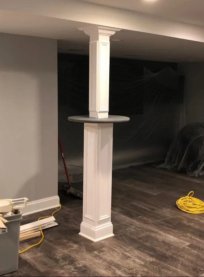 Custom white millwork column with a decorative capital supporting a round bar-height tabletop, built over gray wood-look LVP flooring during a basement build-out