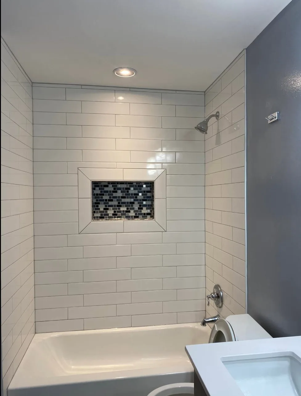 White subway tile tub and shower surround with a framed blue glass mosaic niche, a recessed light, a gray painted accent wall and a white quartz vanity top